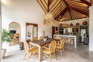 A spacious dining area with a wooden table and wicker chairs, a kitchen with modern appliances, and a large mirror reflecting the room. - Casa Nyang Uluwatu (Pecatu)