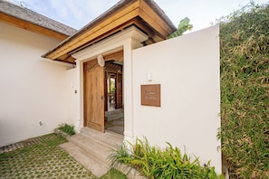 A building entrance with a wooden door, a nameplate, and a small garden area. - Casa Nyang Uluwatu (Pecatu)