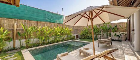 A swimming pool with lounge chairs and an umbrella, surrounded by bamboo fencing and tropical plants.