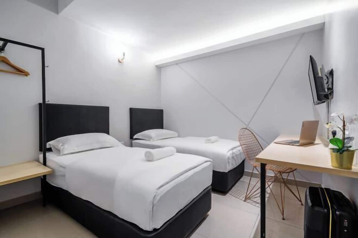 Moderni hotellihuone, jossa on kaksi erillistä sänkyä, työpöytä kannettavalla tietokoneella, tuoli, seinälle asennettu televisio ja seinään kiinnitetty naulakko.