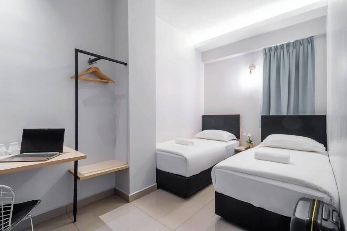 Hotellihuone, jossa on työpöytä, kaksi sänkyä, vaateteline ja pieni yöpöytä, jossa on lamppu.
