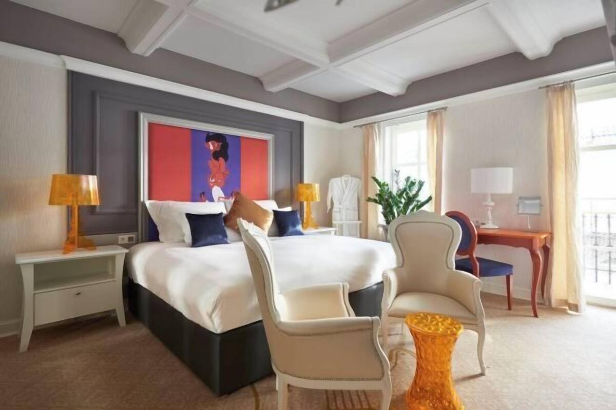 Ett hotellrum med en stor säng, två sängbord, ett skrivbord, två stolar och ett stort fönster med gardiner.