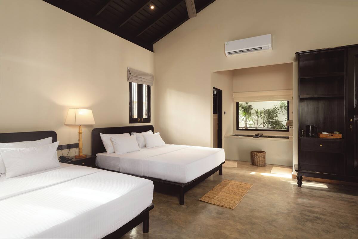 A hotel room with two beds, a lamp, an air conditioner, and a view of greenery through a window.