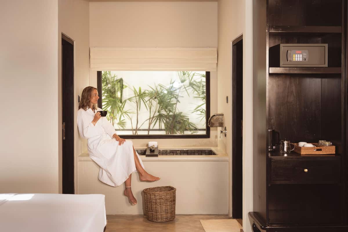 A person in a white robe sitting by a window with a view of palm trees, a sink, and a microwave on a dark wooden cabinet.