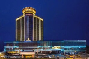 A hotel with a prominent central tower, a large entrance with a covered area, and a parking lot with several cars.