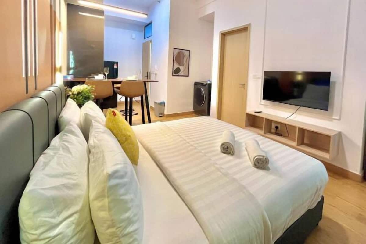 A modern hotel room with a bed, a TV, a desk with a chair, and a small kitchenette area.
