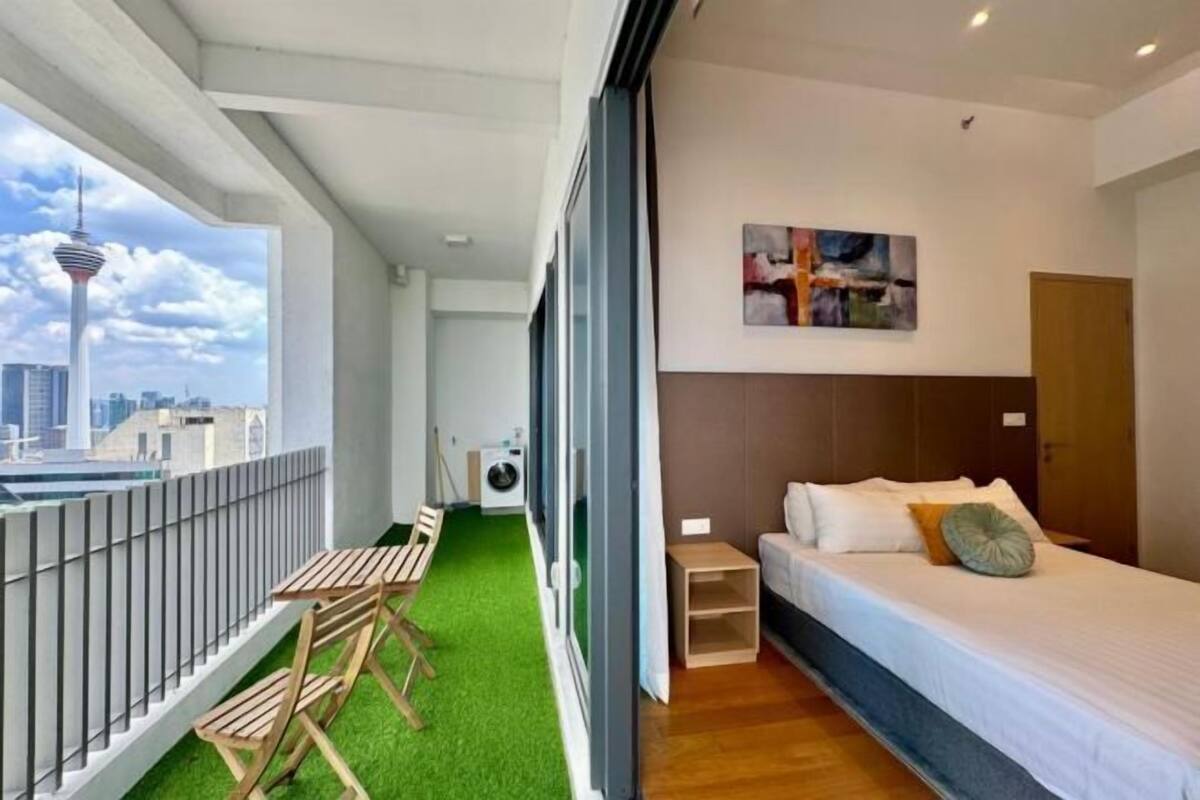 A modern hotel room with a bed, a small table, a chair, a window with a city view, and a wall-mounted artwork.