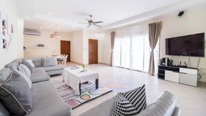 A spacious living room with a grey sectional sofa, a coffee table, and a flat-screen TV on a white cabinet. - Spacious 5-Bed Villa: 7 Mins to Walking Street (Pattaya City)