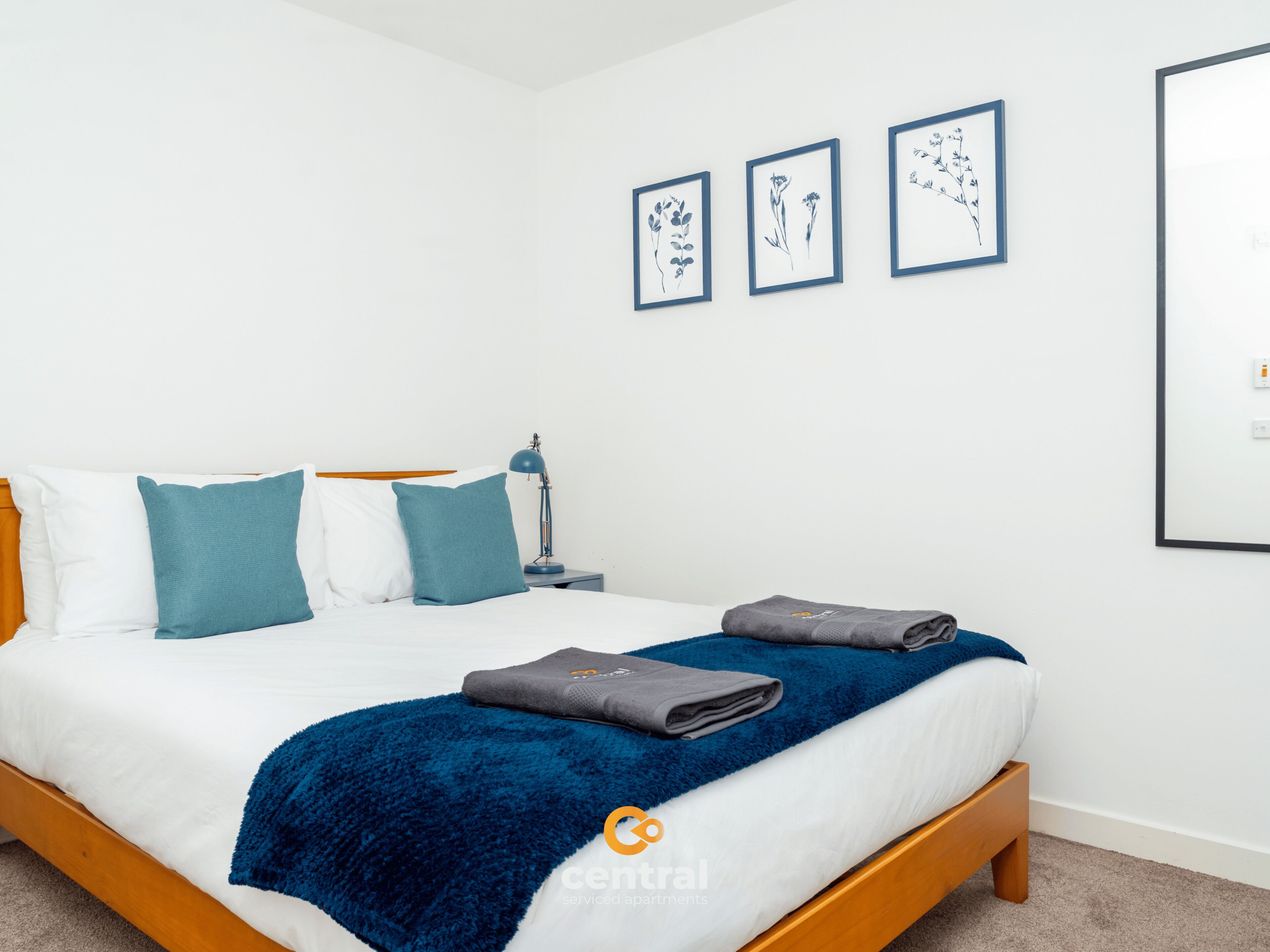 Ein ordentlich gemachtes Bett mit blauer Decke und Kissen, einer Nachttischlampe und drei gerahmten Pflanzenbildern an der Wand.