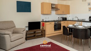 Eine moderne Wohnung mit Wohnzimmer, Küche und Essbereich. Sie umfasst eine Couch, einen Fernseher, Küchengeräte und einen Esstisch mit Stühlen.