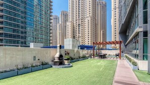 A rooftop garden with a barbecue area, surrounded by high-rise buildings. - Marina Quays Residences - Dubai Marina (Dubai)