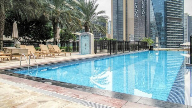 A swimming pool with lounge chairs and palm trees, surrounded by modern high-rise buildings. - Marina Quays Residences - Dubai Marina (Dubai)