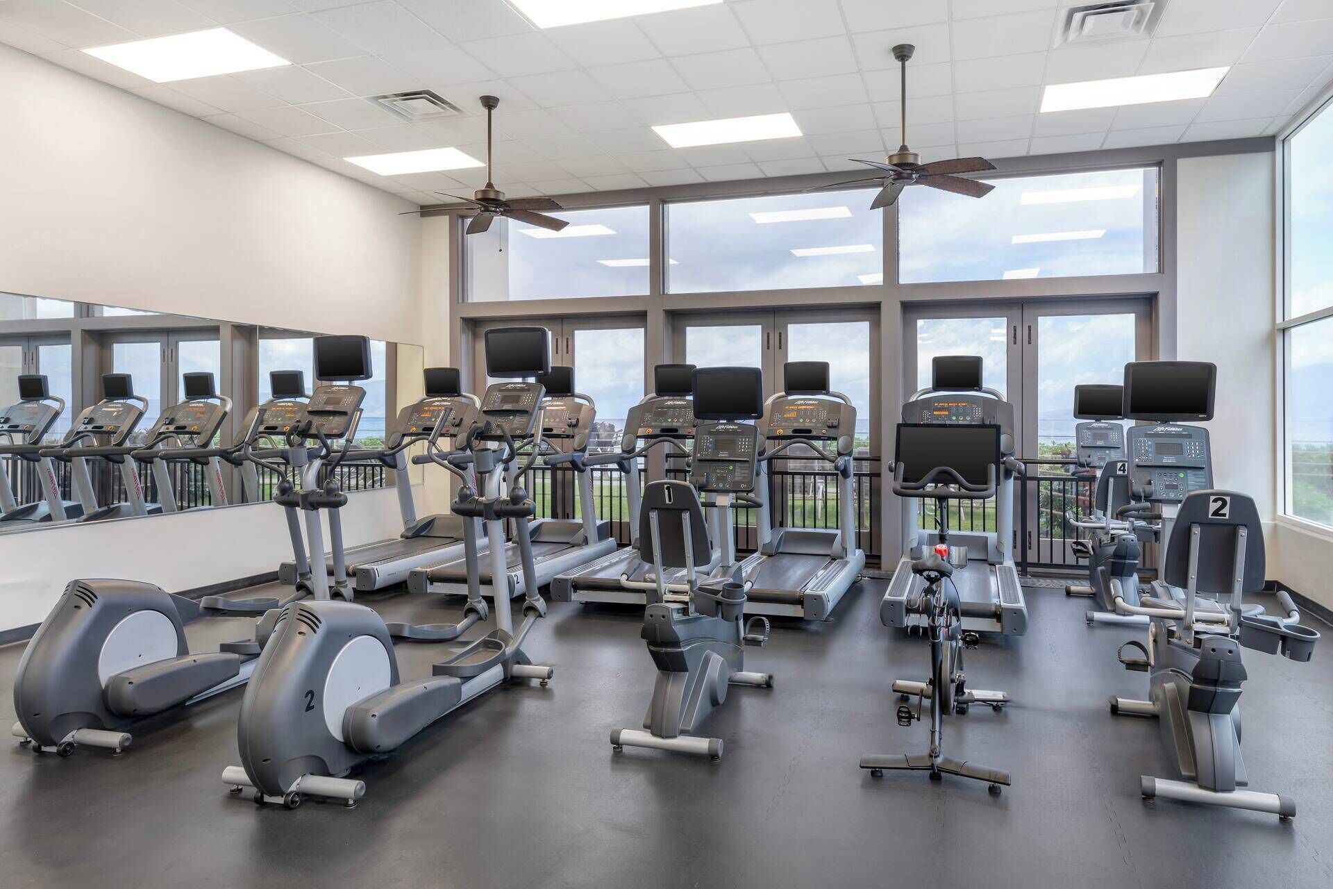 A gym with multiple treadmills, elliptical machines, and a rowing machine, with large windows offering a view of the outdoors.