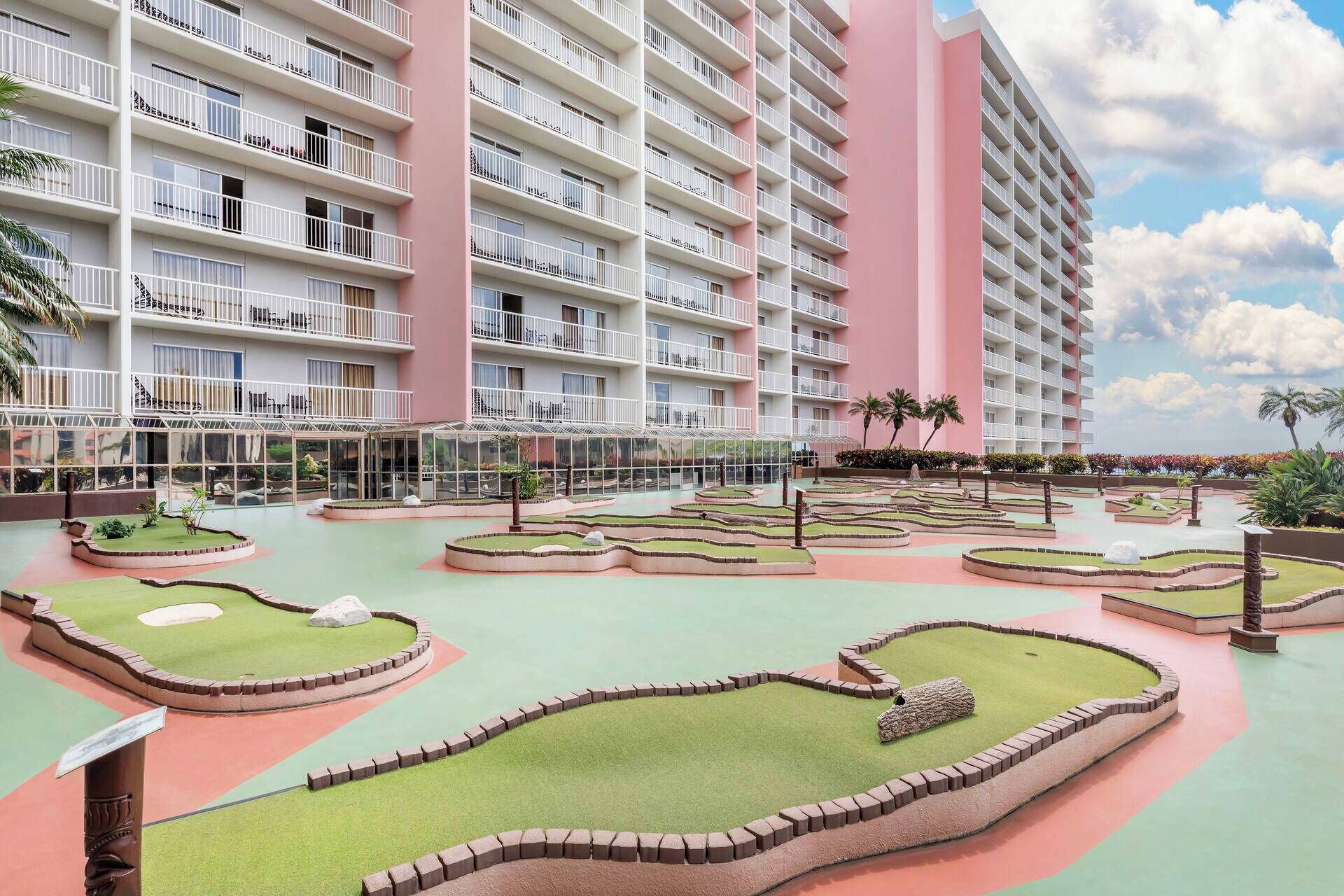 Een hotel met meerdere verdiepingen, met balkons, een aangelegde binnentuin met kunstgras en rotsformaties, en een heldere lucht.