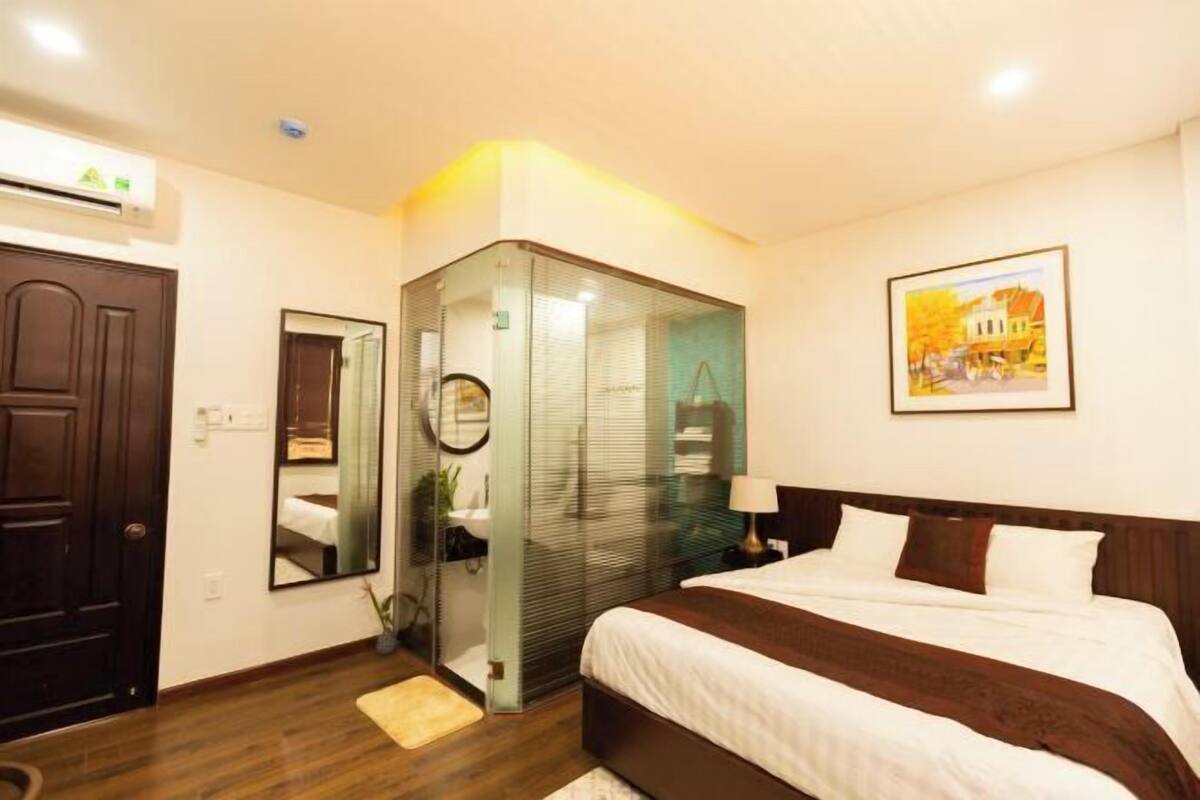 A hotel room with a bed, a wardrobe, a mirror, a painting, and an air conditioning unit.