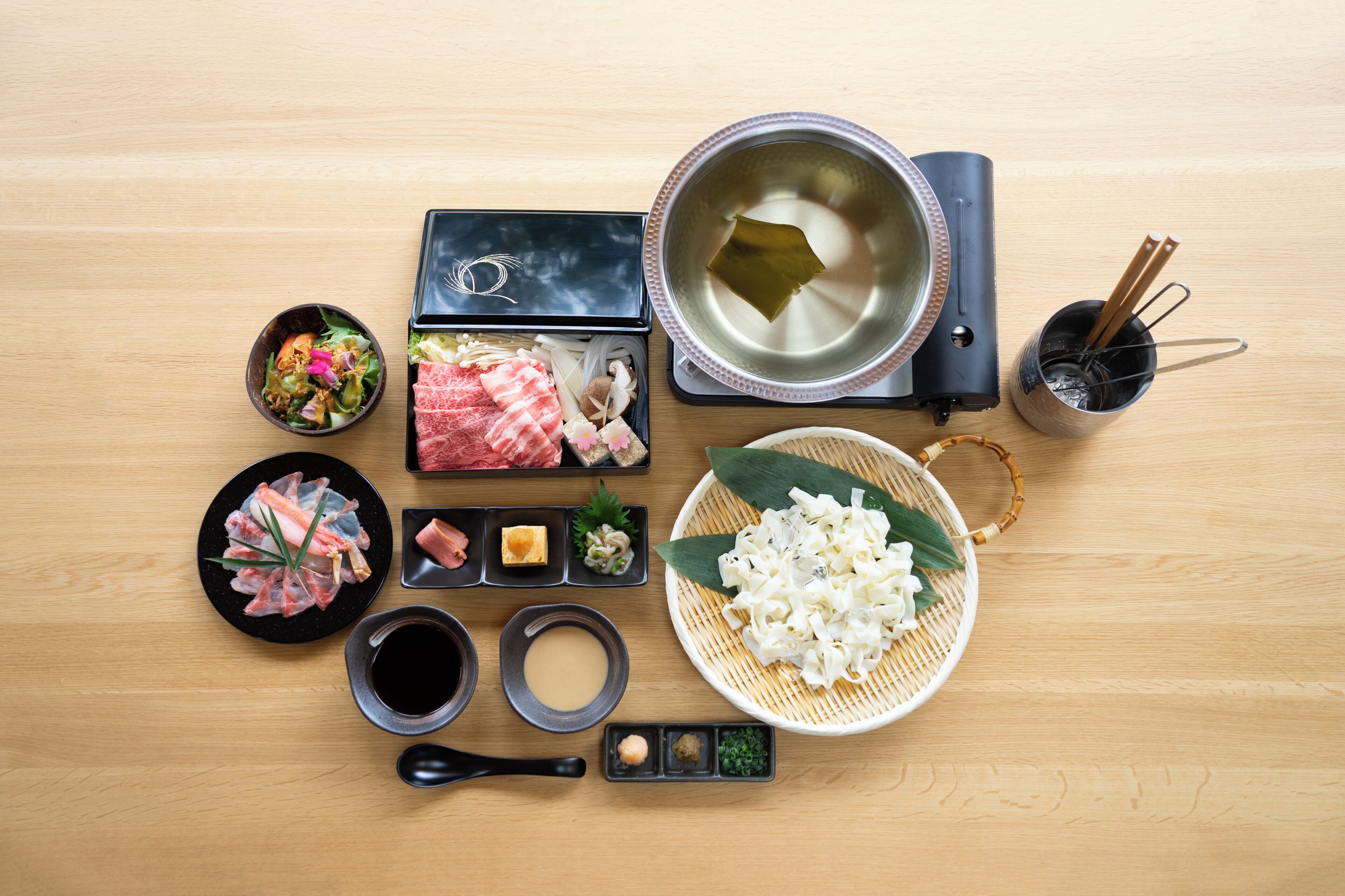 A wooden table with various Japanese dishes, including sashimi, sushi, and a hot pot setup.