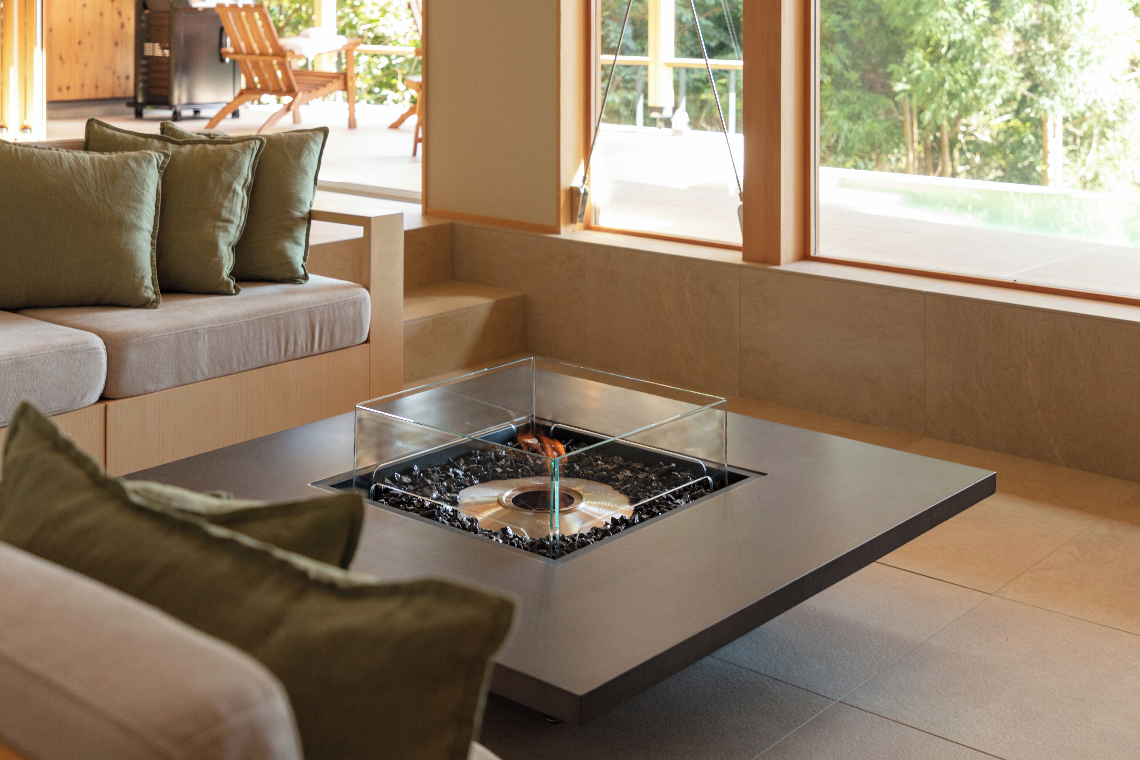 A modern living room with a glass-top coffee table, a sofa with green cushions, and a view of the outdoors through large windows.