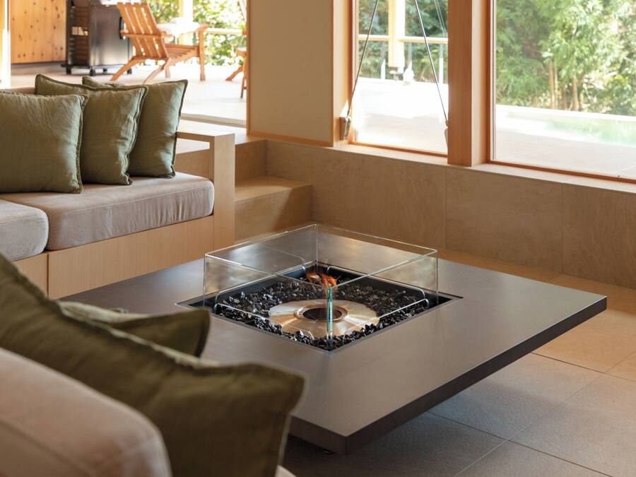 A modern living room with a glass-top coffee table, a sofa with green cushions, and a view of the outdoors through large windows.