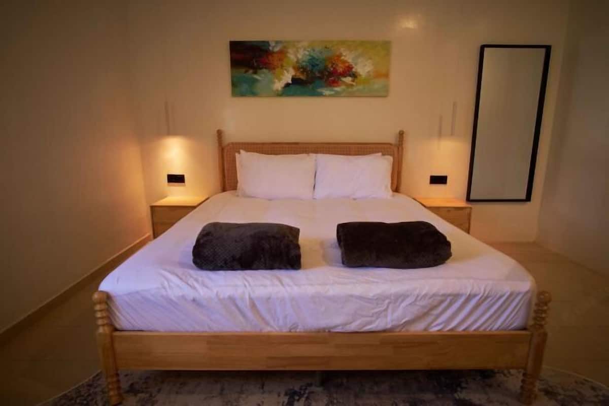 Un letto singolo con biancheria da letto bianca e due cuscini scuri, un quadro al muro e uno specchio a destra.