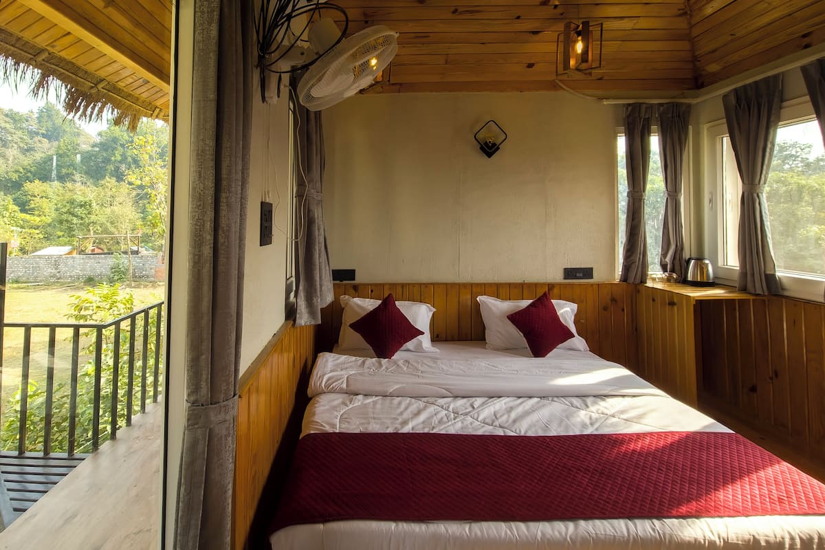 Un dormitorio con cabecera de madera, cama con ropa blanca y detalles rojos, ventana con cortinas y vistas a la vegetación.