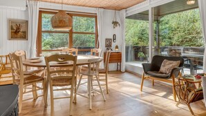 A dining area with a wooden table and chairs, a hanging light fixture, and large windows offering a view of greenery.