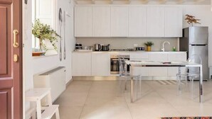 A modern kitchen with white cabinets, a stainless steel refrigerator, and a dining area with a clear table and chairs. - Suite 18 • Loft Incantevole con Balcone sul Mare di Ortigia (Siracusa)
