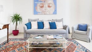A living room with a grey sofa, blue cushions, a glass coffee table, and a patterned rug. - Suite 18 • Loft Incantevole con Balcone sul Mare di Ortigia (Siracusa)