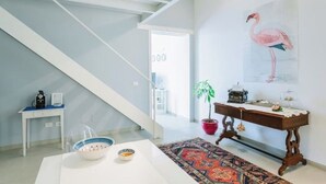 A modern living room with a staircase, a desk, a potted plant, and a painting of a flamingo. - Suite 18 • Loft Incantevole con Balcone sul Mare di Ortigia (Siracusa)