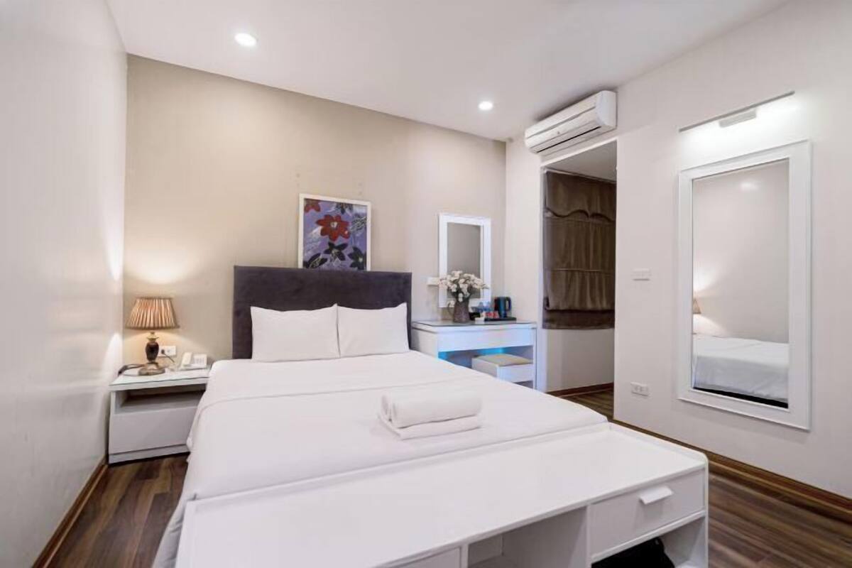 A modern hotel room with a large bed, a bedside table with a lamp, a dresser, a mirror, and an air conditioning unit.