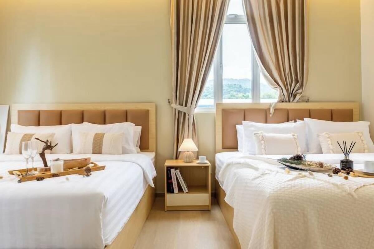 A hotel room with two beds, a nightstand with a lamp, a tray with glasses and a book, and a window with curtains.