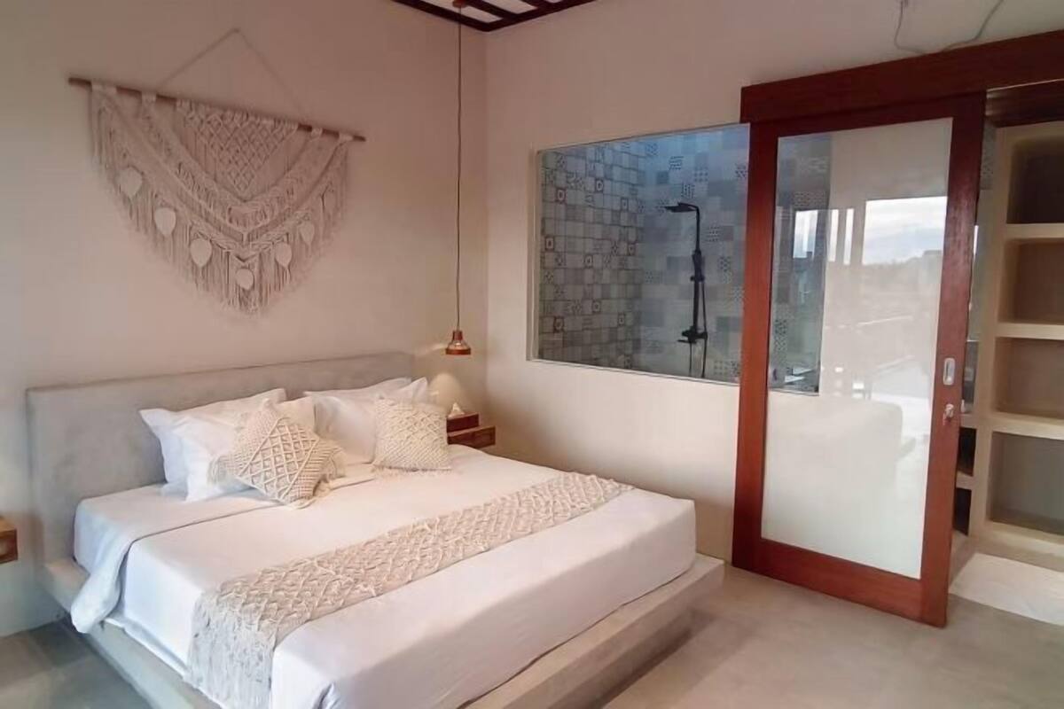A bedroom with a bed, a macrame wall hanging, a large window with a view, and a sliding glass door.