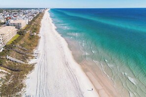 Une vue aérienne d’une plage avec une longue étendue de sable, un océan bleu et limpide, et une zone résidentielle avec des bâtiments et une promenade en bord de mer.