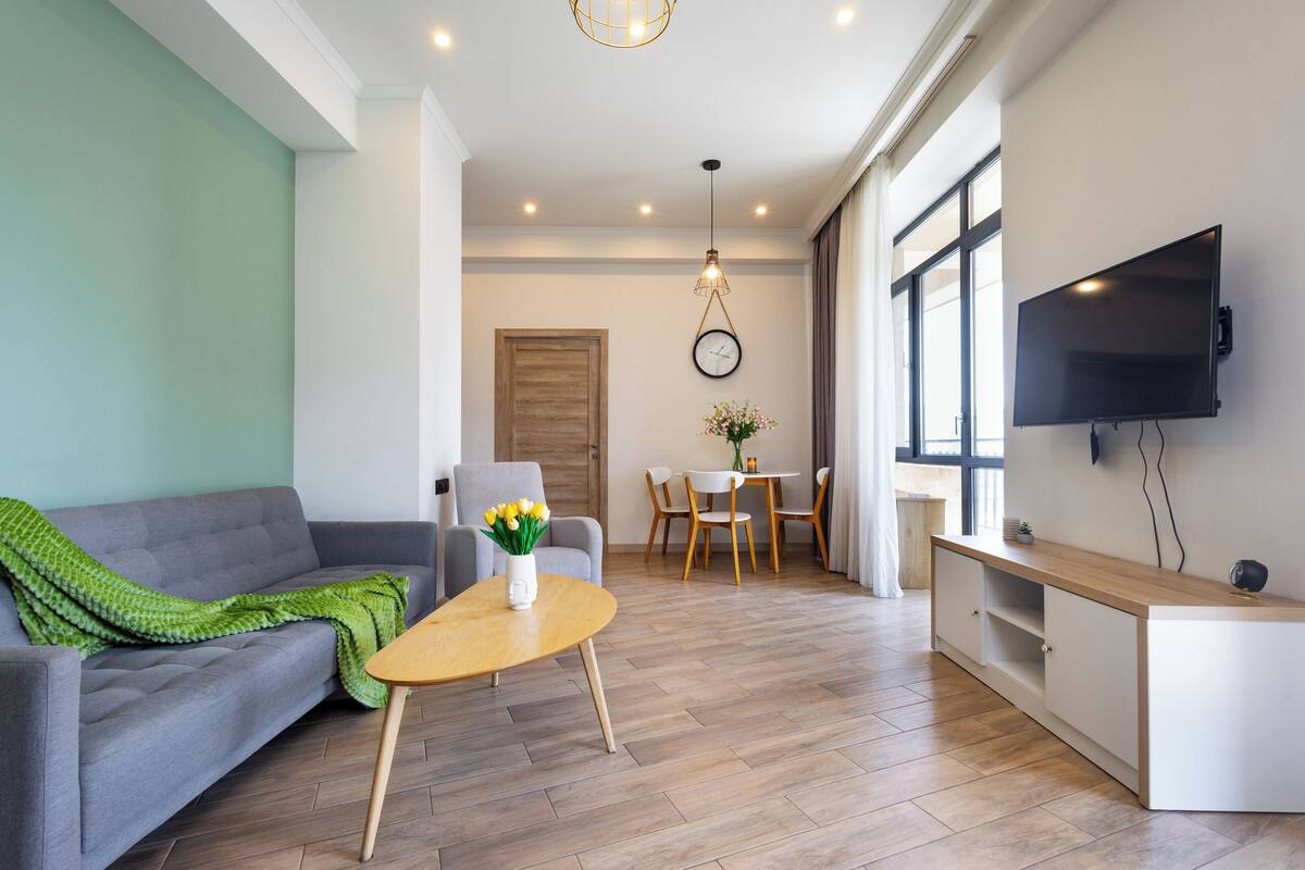 A modern living room with a sofa, a coffee table with flowers, a dining area with chairs, a TV mounted on the wall, and a wooden floor.