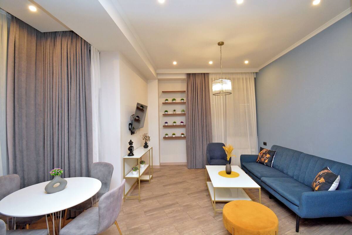 A modern living room with a sofa, a round table, and a coffee table. There are shelves with plants and a TV mounted on the wall.