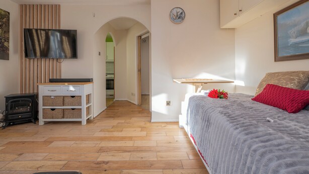 A bedroom with a bed, a TV, a wooden cabinet, a fireplace, and a hallway leading to a kitchen. - Glenville Apartment (Greater London)
