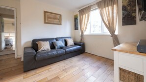 A room with a black leather sofa, wooden flooring, a white cabinet, and a window with curtains. - Glenville Apartment (Greater London)