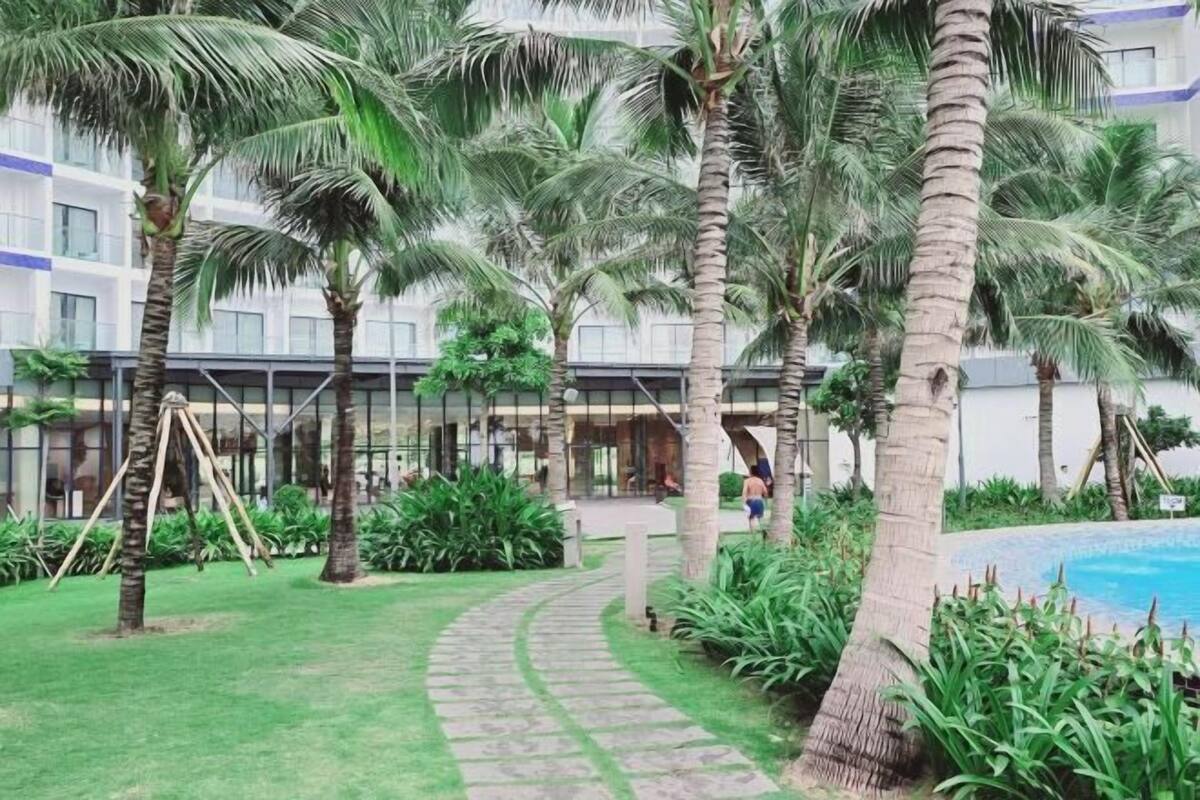 A pathway leading to a pool area surrounded by palm trees and greenery, with a building in the background.