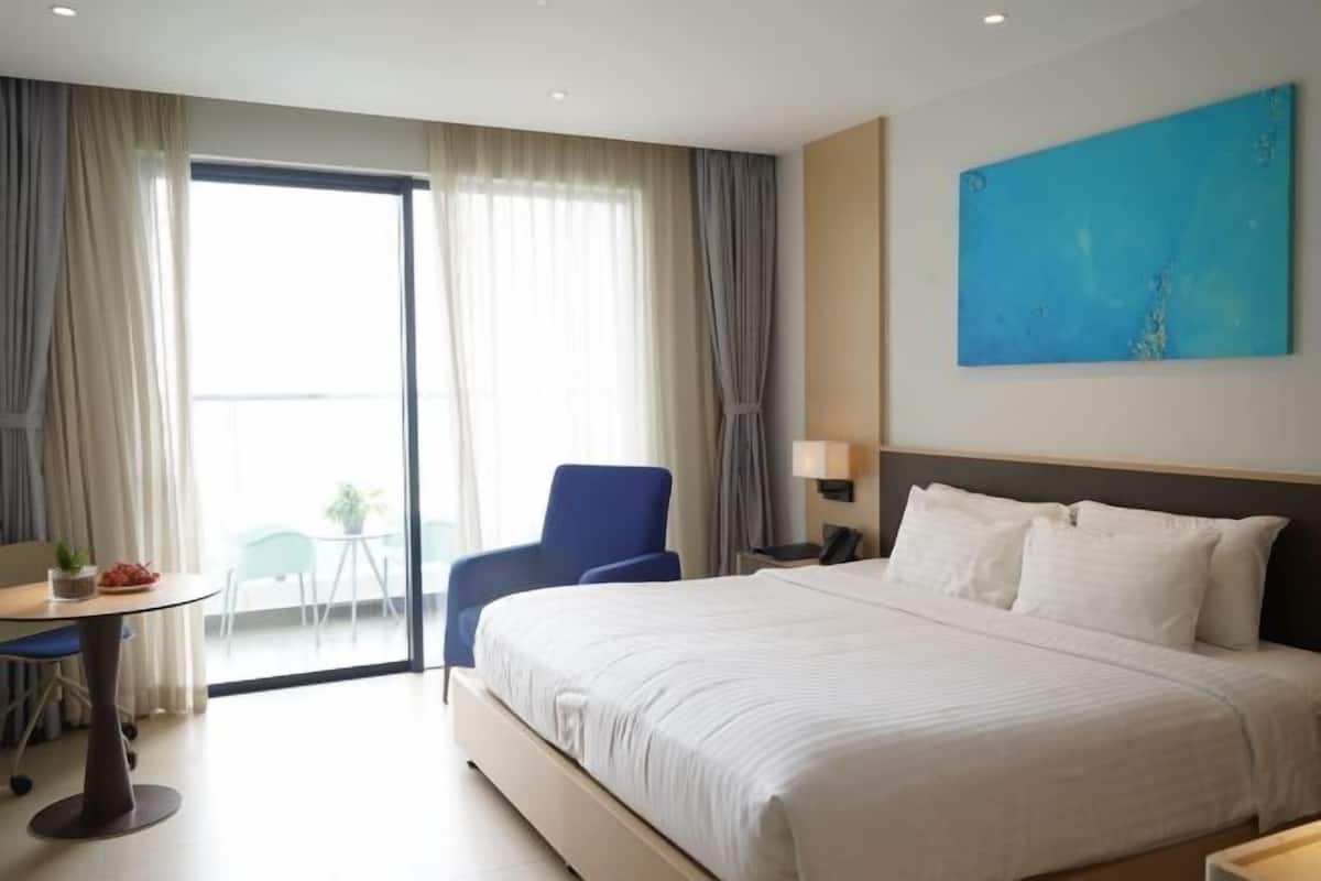 A hotel room with a large bed, a desk with a chair, a small table with a plant, and a view of the outdoors through a sliding door.