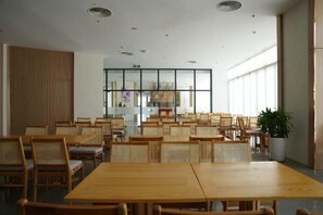 A spacious dining area with multiple wooden tables and chairs, a large window providing natural light, and a potted plant adding greenery. - Signature Beach at The Arena Cam Ranh (Cam Ranh)
