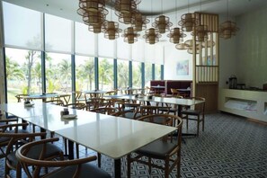 Un comedor amplio con varias mesas y sillas, ventanas grandes que dan a vista de palmeras, y una moderna lámpara de techo.