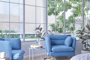 Ein modernes Wohnzimmer mit einem blauen Sofa, einem kleinen runden Tisch mit einer Vase und Obst, sowie großen Fenstern mit Pflanzen.