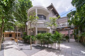 Un patio con un edificio que tiene un balcón curvo, rodeado de árboles y mobiliario exterior.