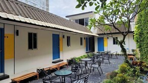A courtyard with outdoor seating, including tables and chairs, surrounded by greenery and a building with a tiled roof. - RYB Hostel Phuket (Phuket)