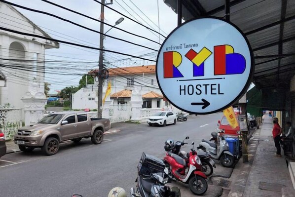 A hostel sign with a directional arrow, parked motorcycles, and a street view with buildings. - RYB Hostel Phuket (Phuket)