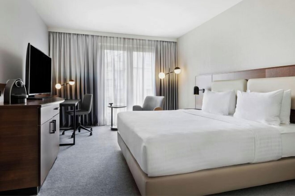 A hotel room with a large bed, a desk with a television, a chair, and a small table.