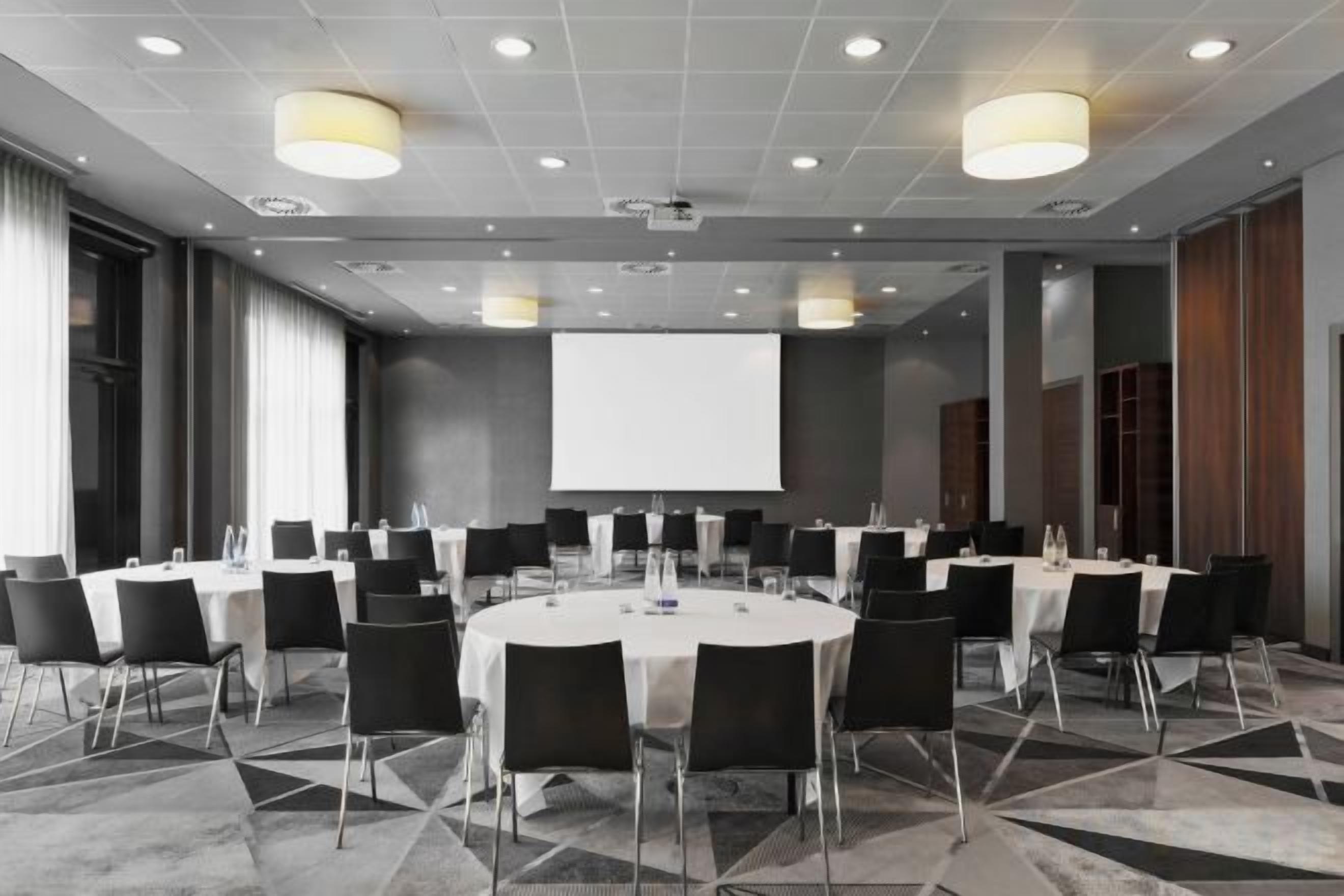 A conference room with round tables, chairs, a projector screen, and a large window.