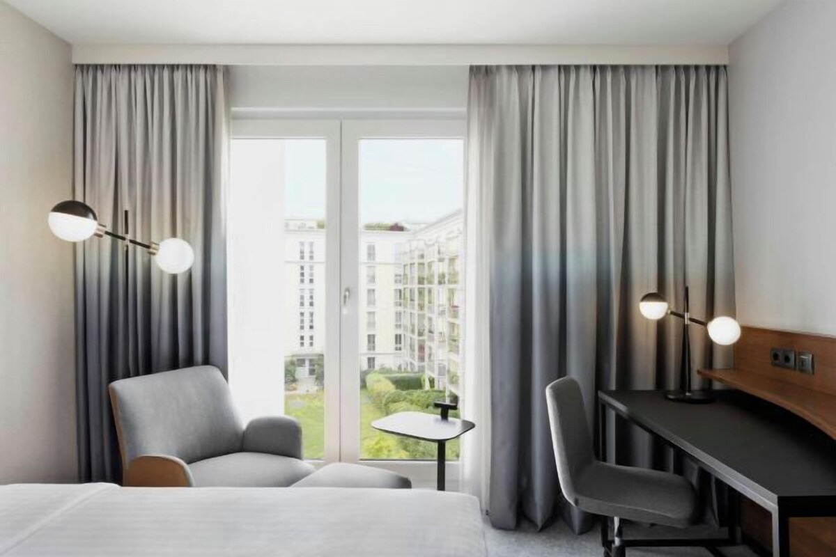 A hotel room with a bed, two chairs, a desk, a small table, and a view of buildings through large windows.