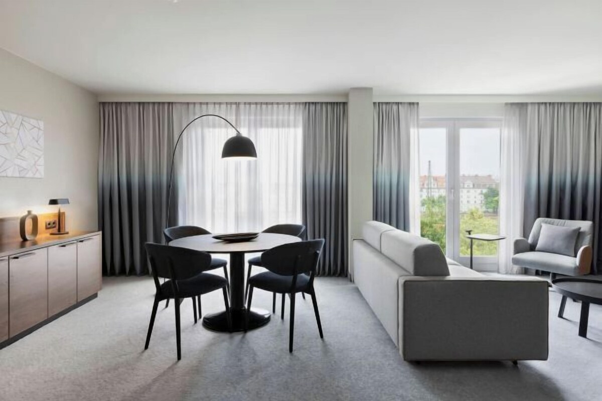 A modern hotel room with a dining area, a sofa, and a view of the city.