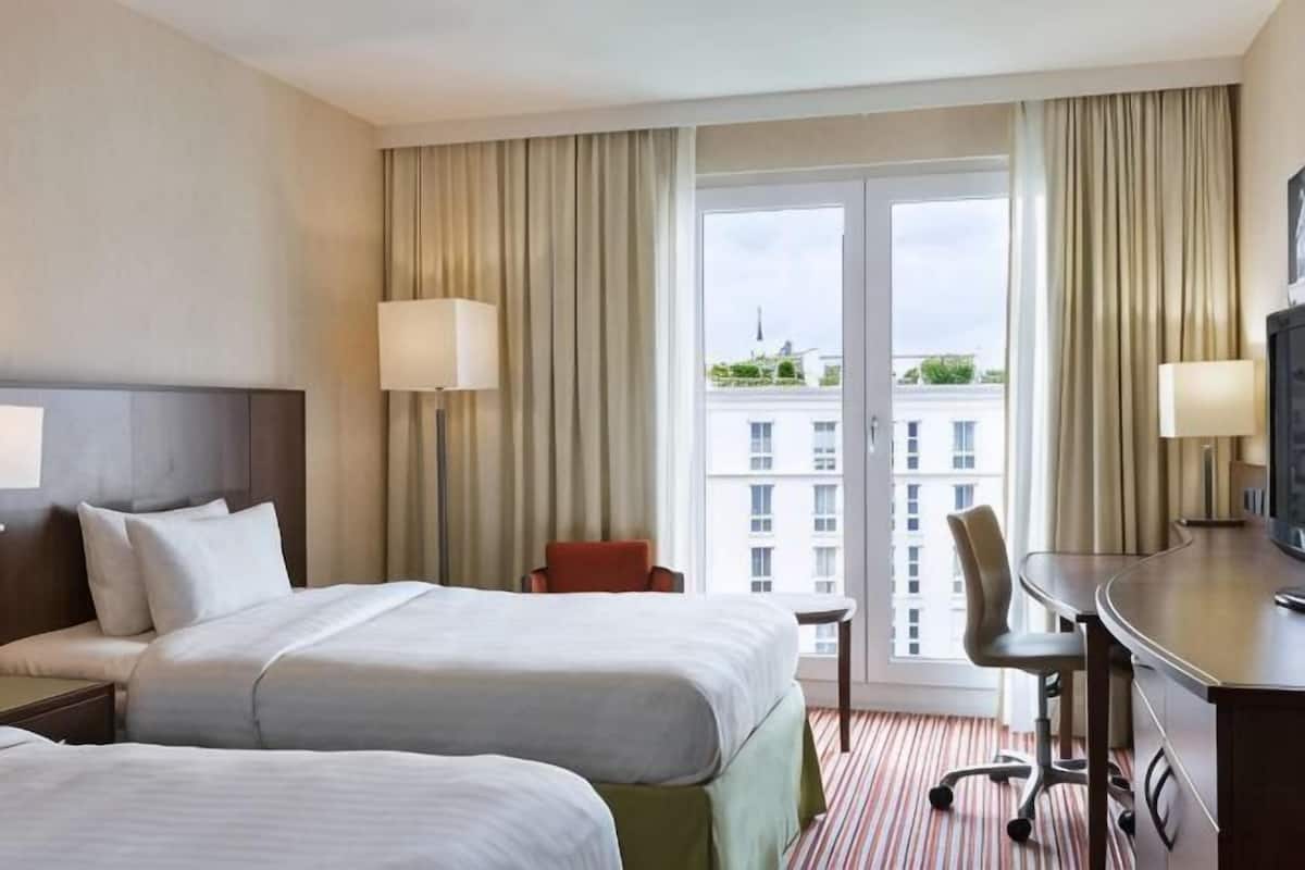 A hotel room with two beds, a desk, a chair, and a large window with curtains.