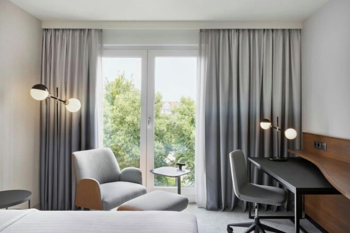 A hotel room with a bed, two chairs, a desk, a small table, and a view through a large window with curtains.